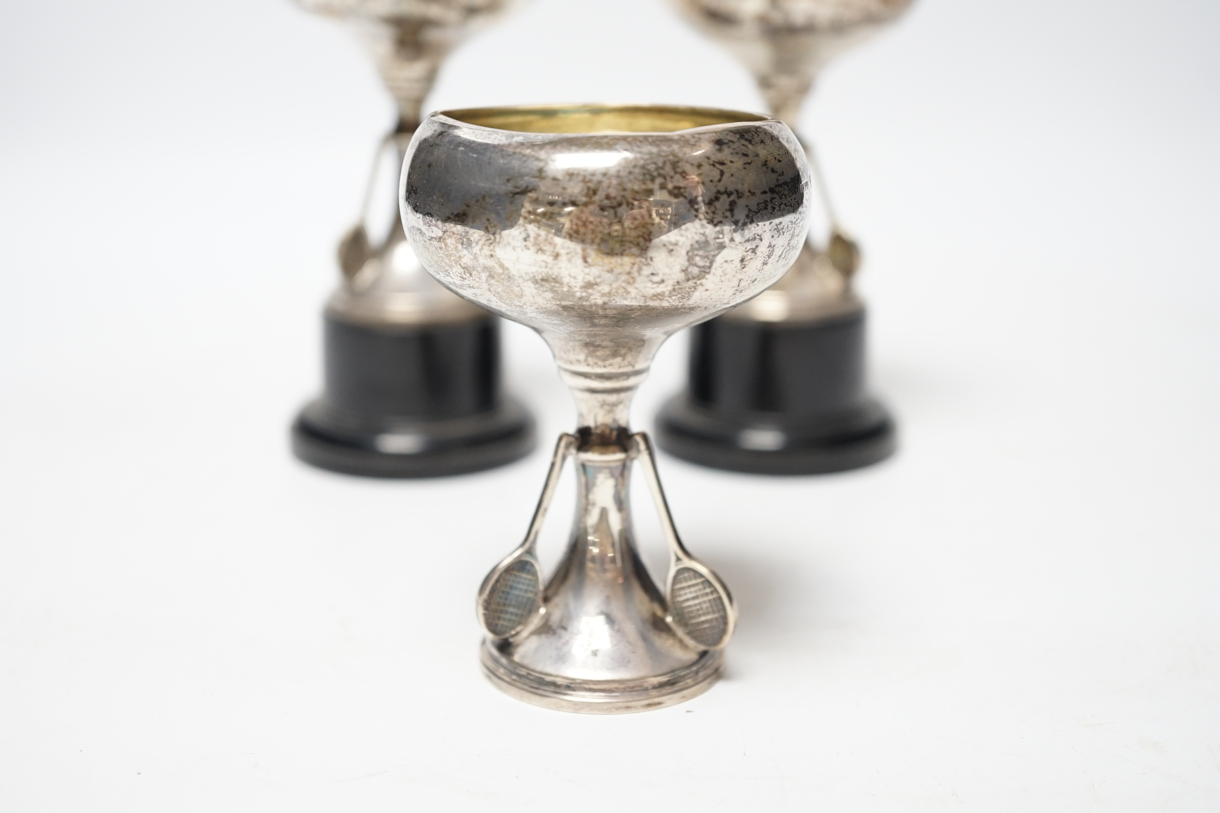 A set of three small George V silver tennis related trophy cups, with racket supports and engraved inscriptions, E & J Leek, 92mm, two with plinth bases.